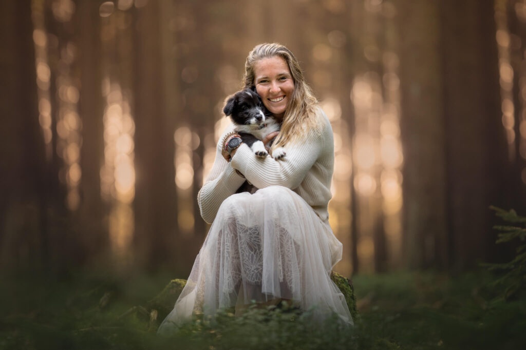 A woman sitting in a sunlit forest, smiling and holding a small black and white puppy. She wears a white sweater and a long, light-colored skirt.