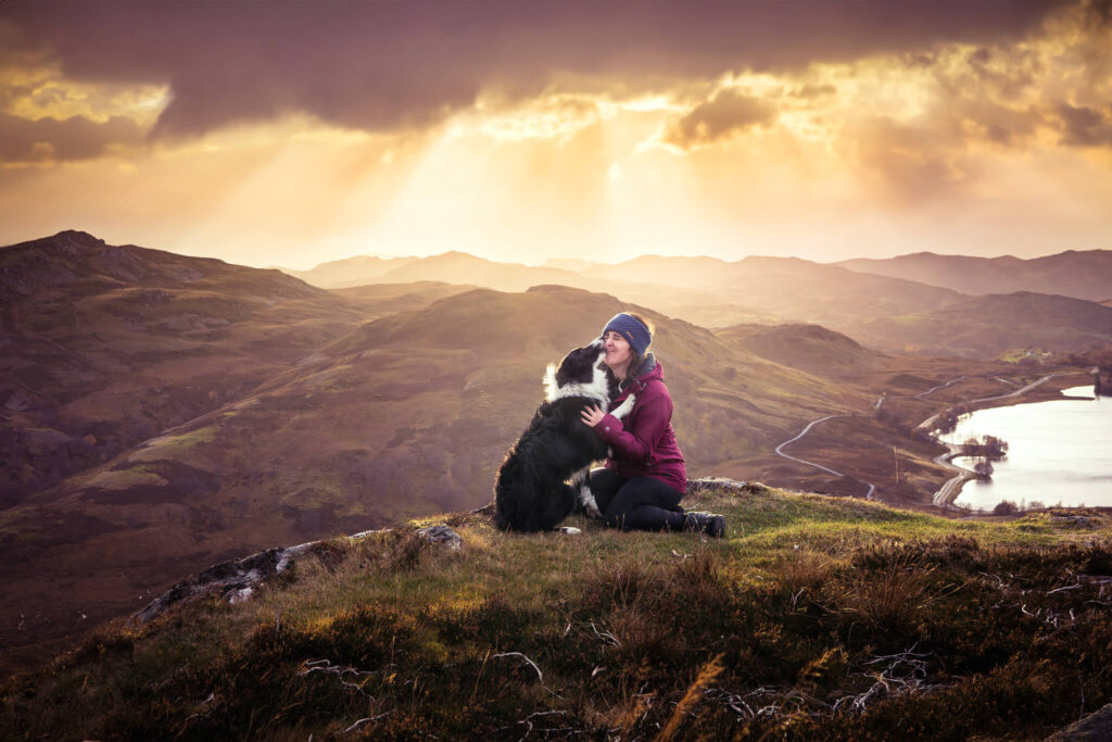 A person in a red jacket kneels and embraces a black and white dog on a grassy hill with rolling hills and a lake in the background under a cloudy, sunlit sky.