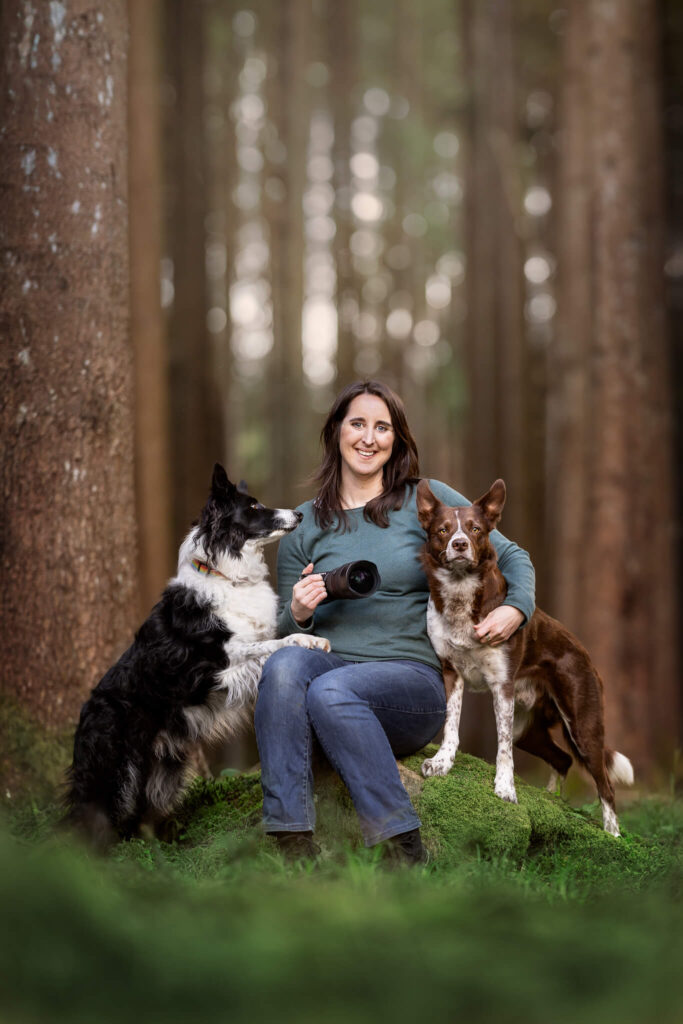 A woman in a green sweater and jeans sits on a rock in a forest, holding a camera lens. She is accompanied by two dogs, one black and white, and the other brown and white.
